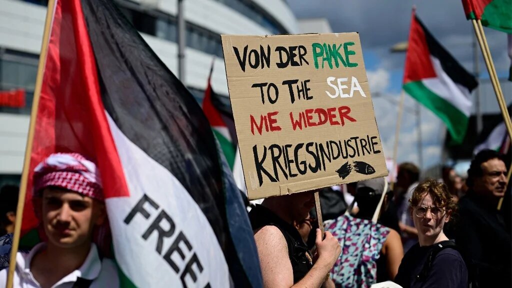 A demonstrator holds a placard reading “From the Panke [River] to the Sea - never again war industry”, as others hold Palestinian flags, in Berlin, Germany, on 11 July 2026 (Tobias Schwarz/AFP)