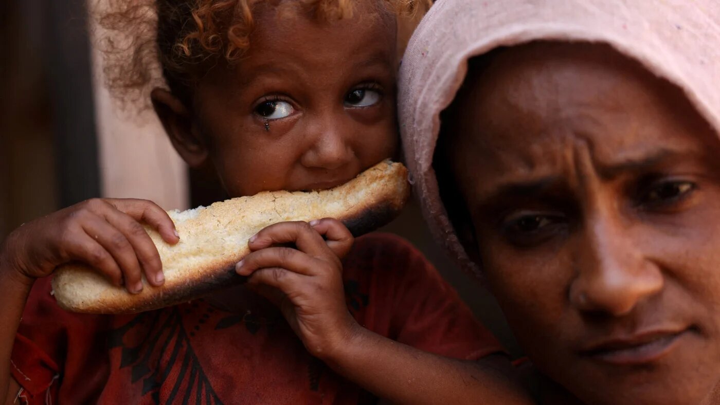 A girl, with her mother, bites on a piece of bread at a camp for displaced people in the city of al-Khokha, Yemen, 25 October 2024 (Reuters/Amr Alfiky)