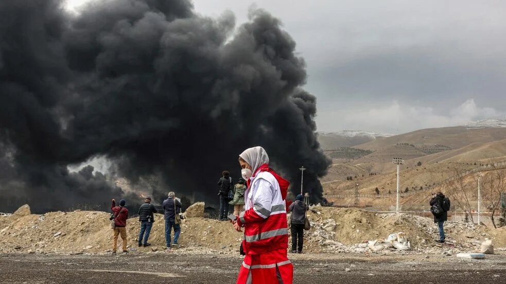 Members of Iran’s Red Crescent society walk near smoke plumes after an air strike on the Shahran oil refinery in northwestern Tehran on 8 March 2026 (AFP)