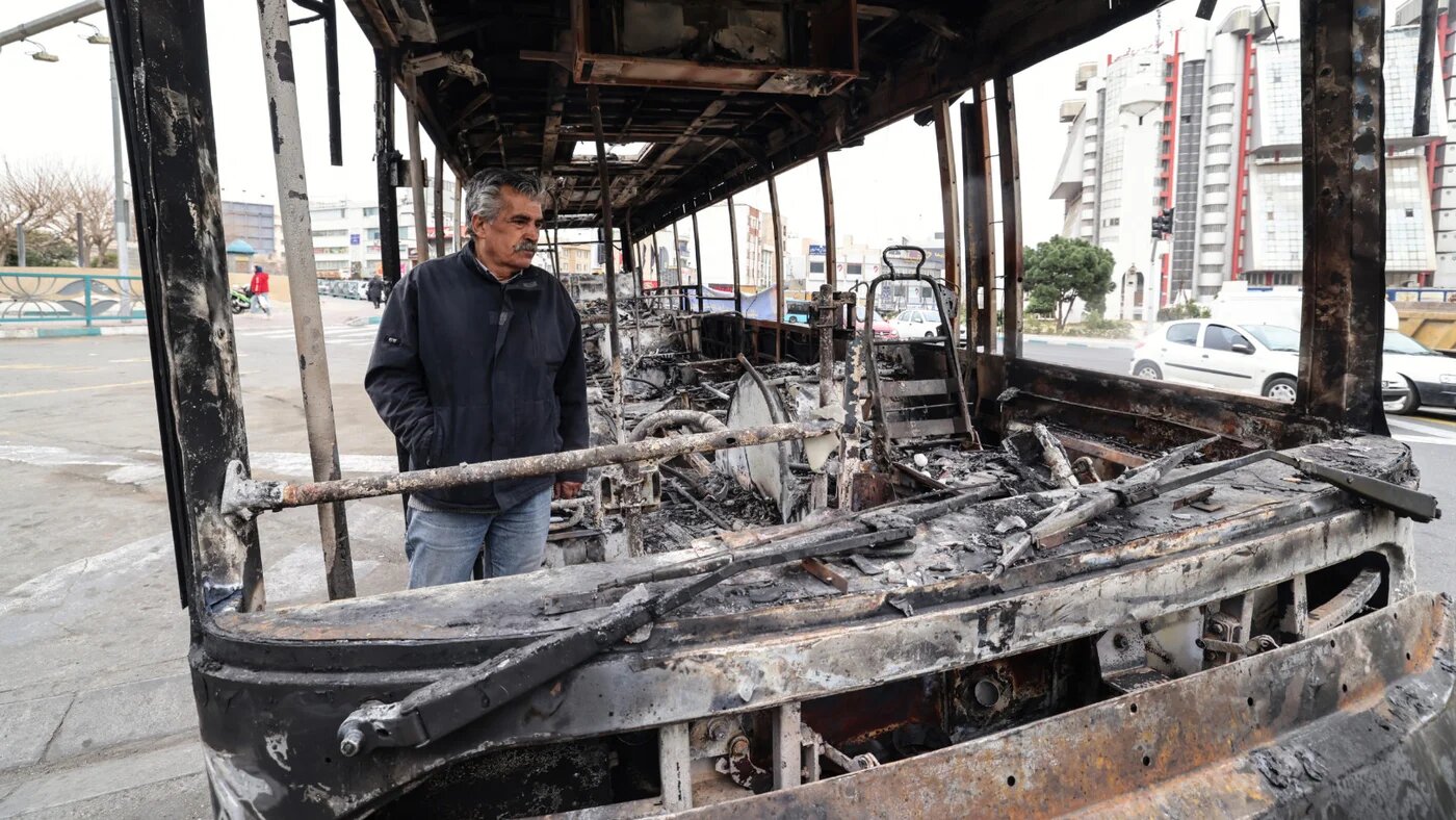 A man stands by the wreckage of a burnt bus bearing a banner (unseen) that reads "This was one of Tehran’s new buses that was paid for with the money of the people’s taxes,” in Tehran's Sadeghieh Square on 15 January 2026 (AFP)