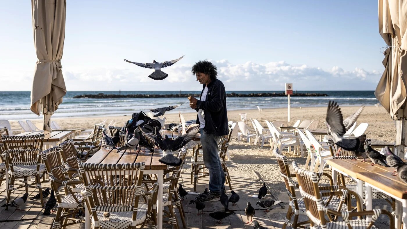 A man feeds pigeons along an empty beach in Tel Aviv on 28 February 2026 (John Wessels/AFP)