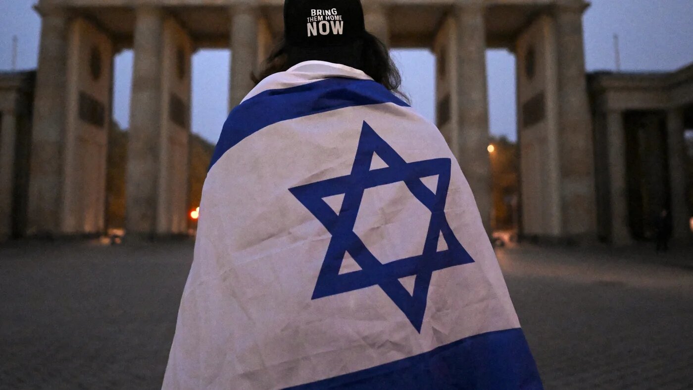 A woman wrapped in an Israeli flag in front of the Brandenburg Gate in Berlin early in the morning of 7 October 2025 (AFP/Tobias Schwarz)