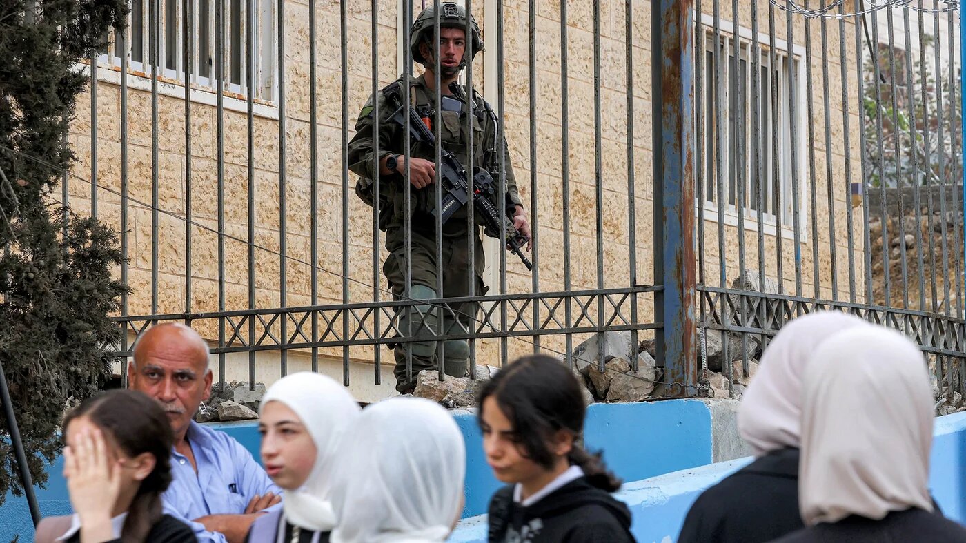 An Israeli army soldier stands from behind a fence as Palestinian schoolgirls make their way to school on the first day of term in Hebron, occupied West Bank on 8 September, 2025 (AFP/Hazem Bader)