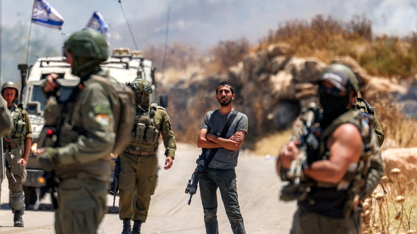 An armed Israeli settler stands between Israeli forces along a road after settlers set fire to an area in the Palestinian village of Idna in occupied West Bank, 5 June 2026 (Hazem Bader/AFP)