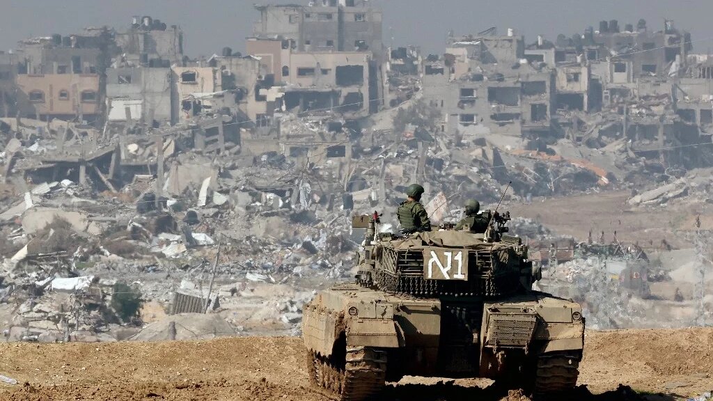 A picture taken from a position in southern Israel along the border with the Gaza Strip shows an Israeli tank rolling along the fence as damaged buildings are see in the Gaza strip on 19 January 2024 (AFP)