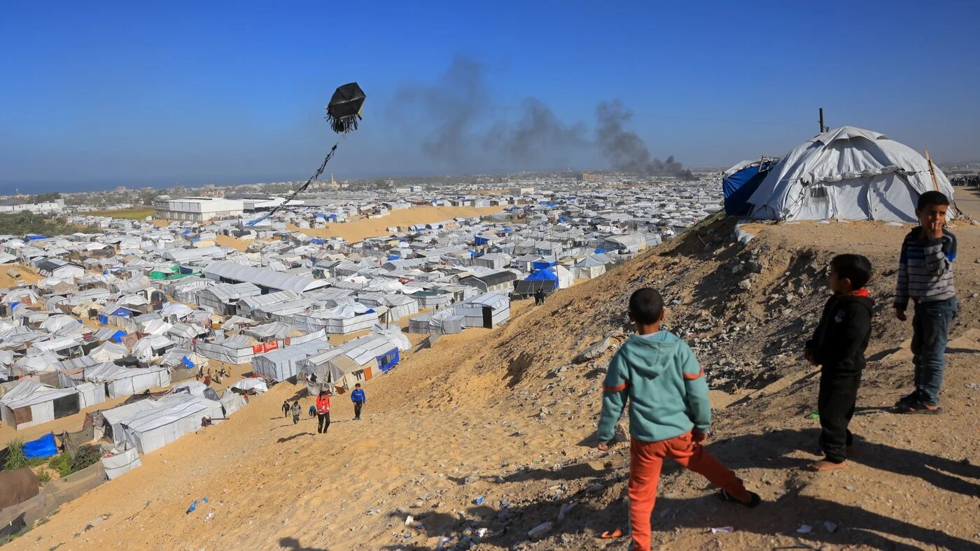 Boys fly a kite against the backdrop of smoke rising from a shelter that houses displaced Palestinians, after an Israeli air strike in the west of Khan Yunis, southern Gaza, 31 January 2026 (Bashar Taleb / AFP)