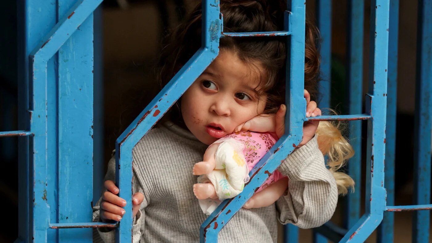 A displaced child from southern Lebanon holds a doll as she looks through a fence at a school-turned shelter, near Sidon, Lebanon, 17 May 2026 (Aziz Taher/Reuters)