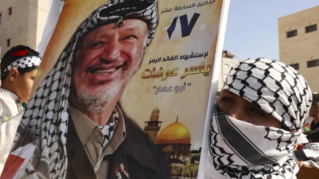 Palestinian supporters of the Fath movement raise a poster of the late leader Yasser Arafat during a rally marking the 17th anniversary of his passing, in the village of Halhul, north the West Bank town of Hebron, on 11 November 2021 (AFP)