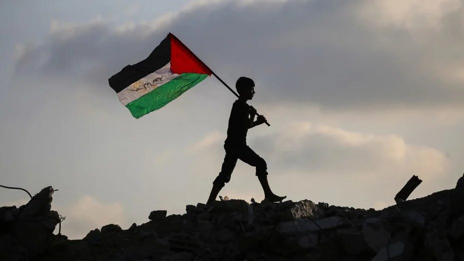 A displaced child waves a Palestinian national flag as he walks on the rubble of a destroyed building in Gaza’s Bureij camp on 22 September 2025 (Eyad Baba/AFP)