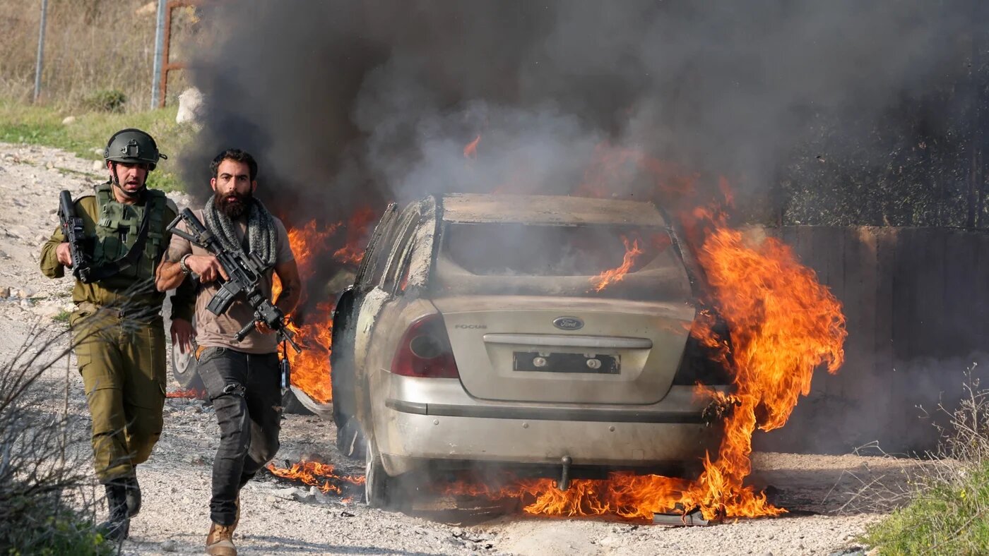 An Israeli troop and settler security officer walk past a burning car reportedly set on fire by settlers in the Palestinian Burin village in the occupied West Bank on 25 February 2023 (AFP)