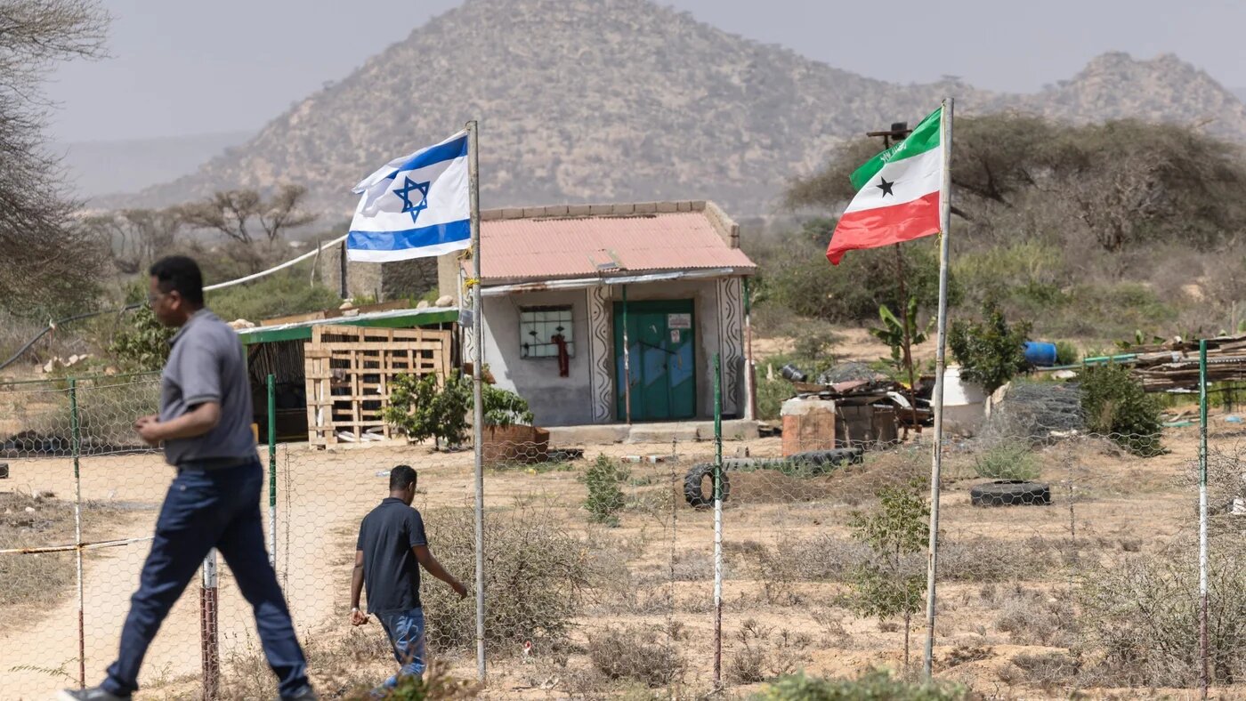 People walk by the flags of Israel and Somaliland flying alongside each other at the entrance to a fruit farm between the capital city of Hargeisa and Port city of Berbera on 19 February 2026 (AFP/Tony Karumba)