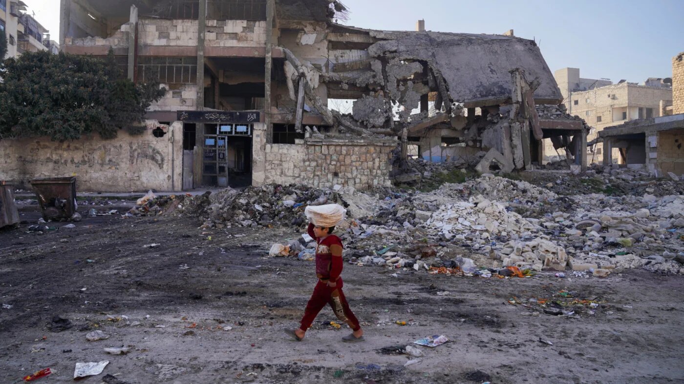 A boy carrying stacks of bread on his head walks past a damaged school in Aleppo, Syria on 21 January 2025 (Karam al-Masri/Reuters)