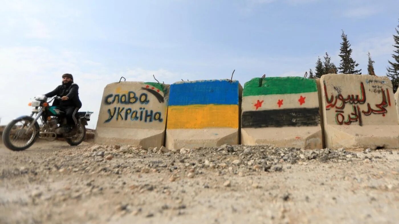 Un homme passe à moto devant des blocs de ciment où sont peints les drapeaux de l’Ukraine et de l’opposition syrienne, ainsi que les slogans « Gloire à l’Ukraine » et « Gloire à la Syrie libre », dans la province d’Alep, le 28 février 2022 (AFP)