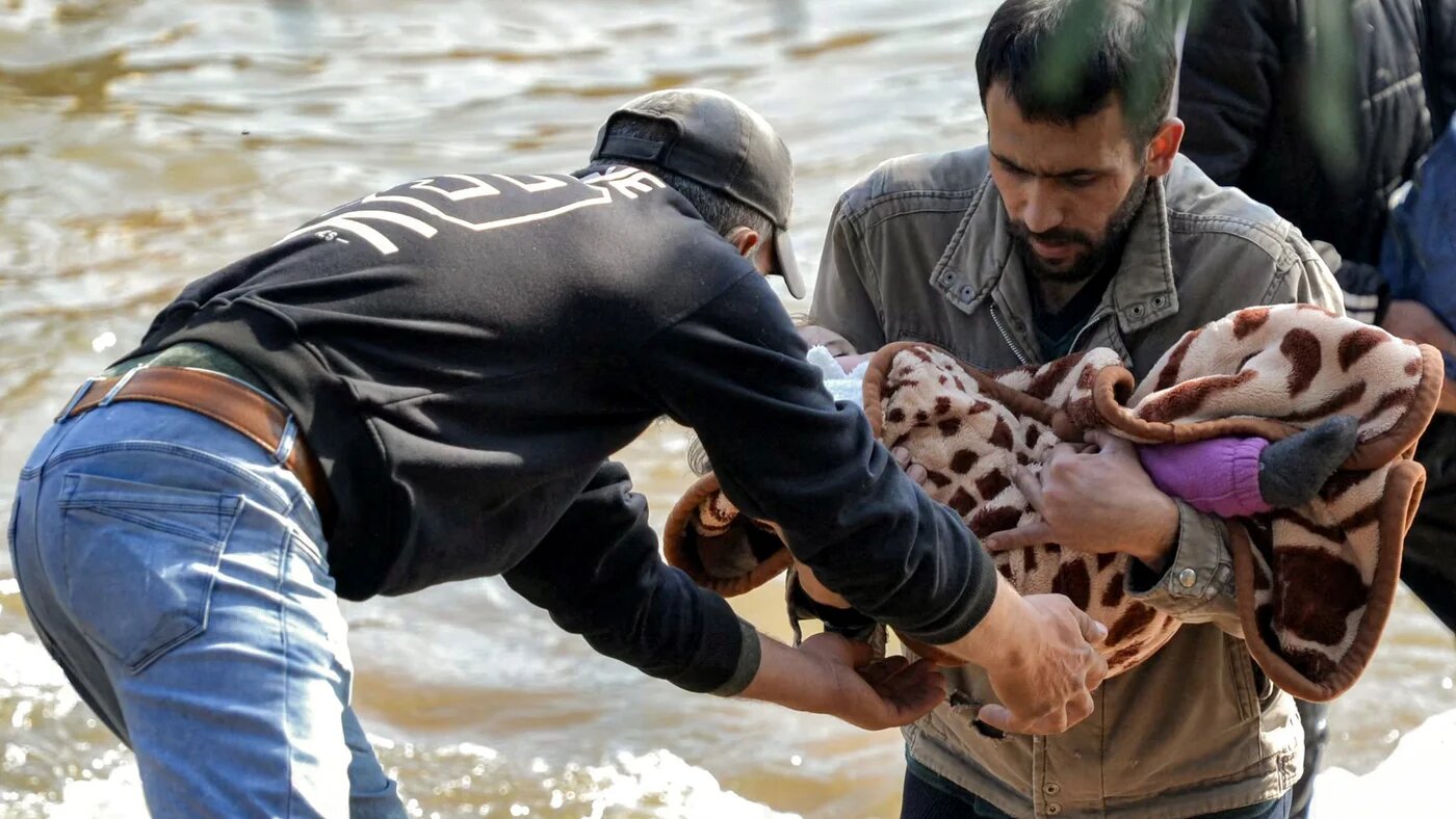 A man receives a blanketed child from another crossing the Nahr al-Kabir river, forming the border between Syria's western coastal province and northern Lebanon in the Hekr al-Daher area on 11 March 2025 (AFP)