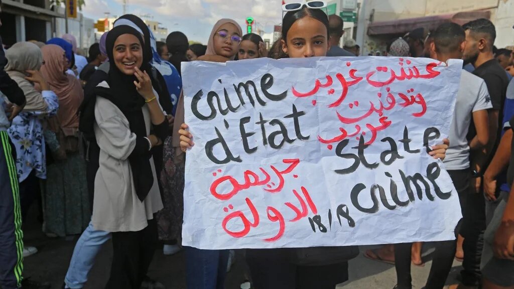 Une femme brandit une pancarte indiquant « J’ai vécu en étranger et j’ai été enterré en étranger » et « crime d’État », alors que les habitants de la ville côtière de Zarzis, dans le Sud-Est de la Tunisie, se rassemblent le 18 octobre lors d’une grève générale afin de réclamer une nouvelle recherche des proches portés disparus lors d’une tentative de migration clandestine en septembre (Fathi Nasri)