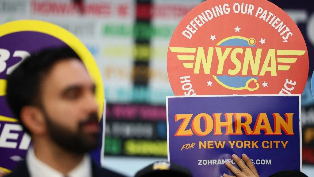 People hold signs as Zohran Mamdani, then a candidate for mayor, speaks during a news conference in New York City on 2 July 2025 (Michael M Santiago/Getty Images/AFP)