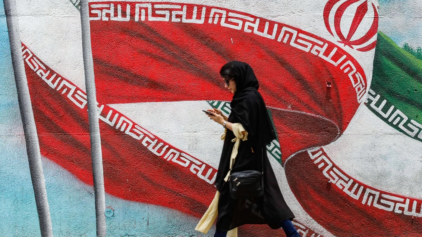 An Iranian girl walks next to a wall painting in Tehran of Iran's national flag while using her phone