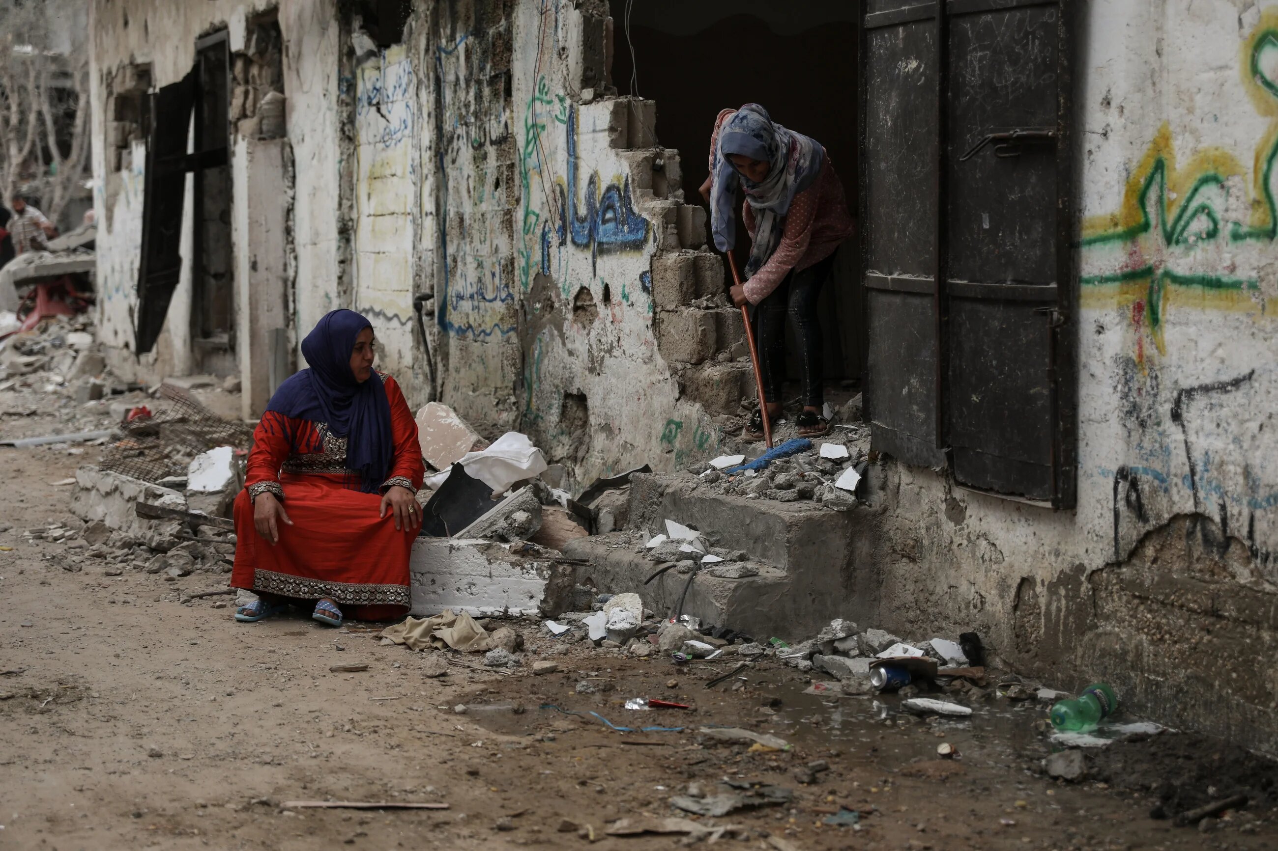 A woman cleans away the rubble and debris from her home, as another woman looks on while sitting on a large piece of building wall ripped from the building's structure