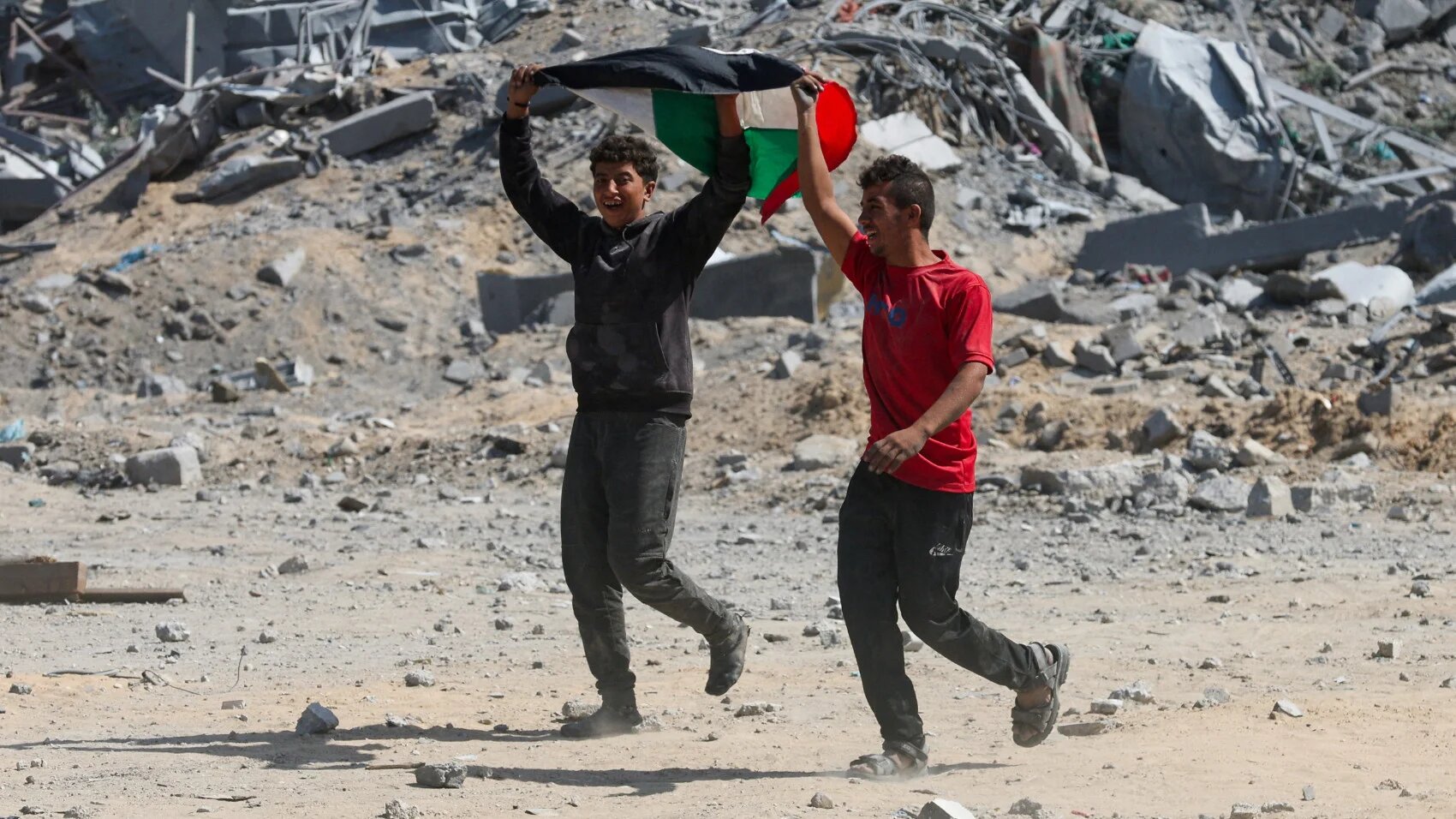Palestinians wave their flag as they walk among the rubble following the withdrawal of Israeli forces from the area, in Khan Younis, in the southern Gaza Strip, 10 October 2025 (Ramadan Abed/Reuters)