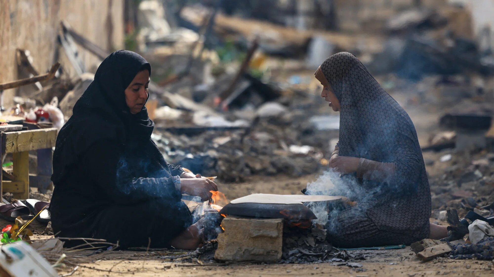Palestinian women bake on wood fire outside their damaged homes in the Khezaa district on the outskirts of the southern Gaza Strip city of Khan Yunis on 25 November 2023 (AFP)