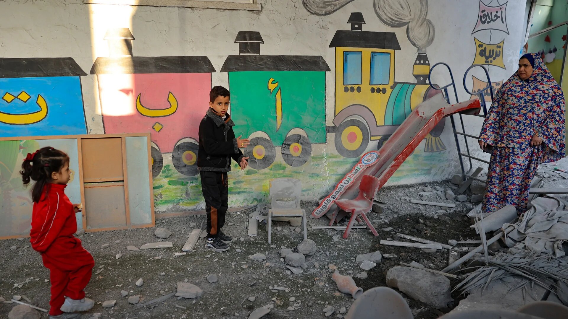A Palestinian woman and children stand amid the rubble at a kindergarten hit by Israeli air strikes in Rafah, in the southern Gaza Strip, on 9 December 2023 (Said Khatib/AFP)