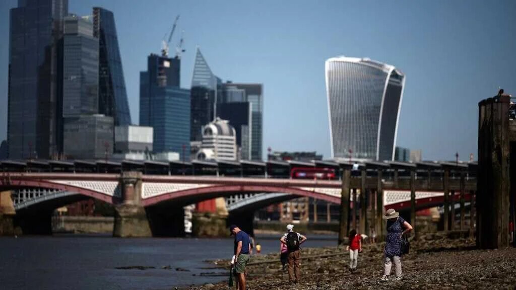 People walk on the shoreline of the River Thames, enjoying the sun and hot weather, backdropped by the City of London skyline, on July 29, 2024.
