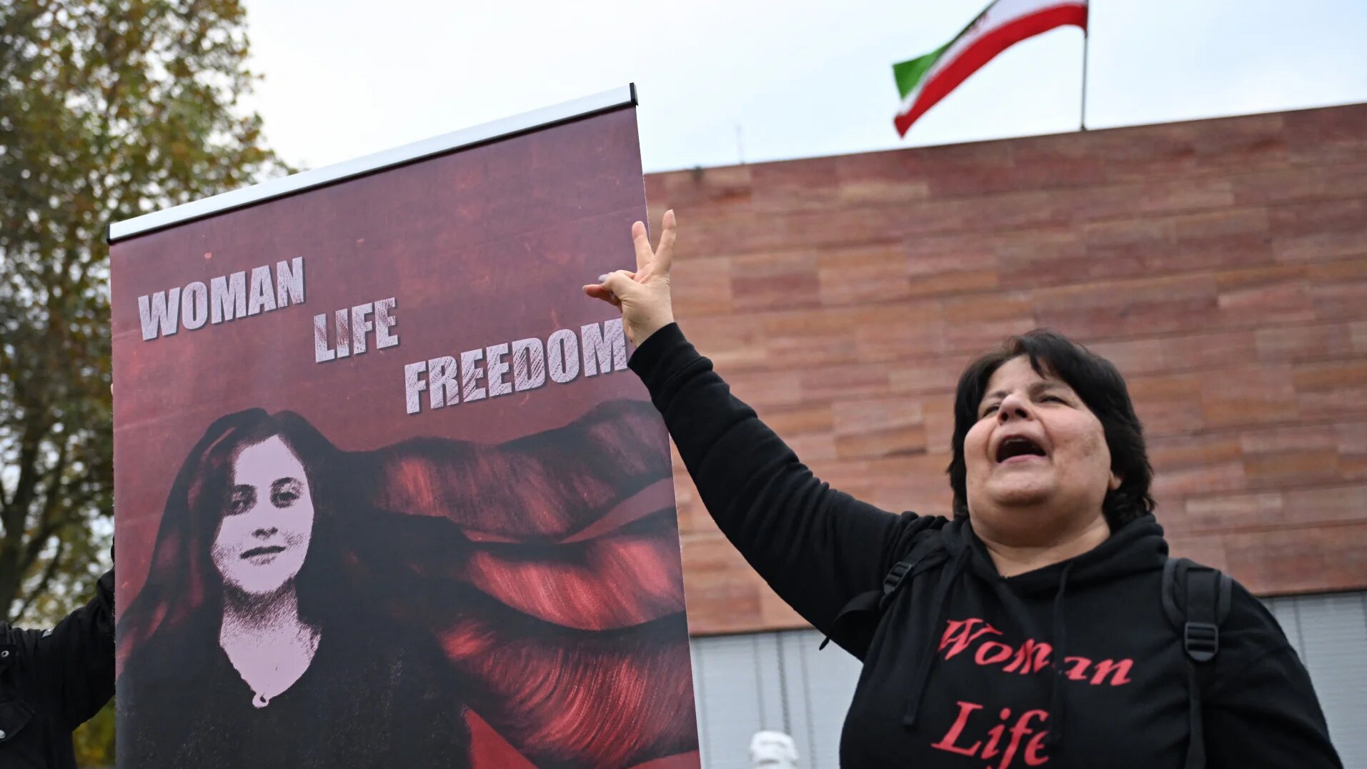 A demonstrator flashes the victory sign next to a placard of Mahsa Amini and the slogan ‘Woman, Life, Freedom’ outside Iran’s consulate in Frankfurt, Germany, on 31 October 2024 (Kirill Kudryavtsev/AFP)