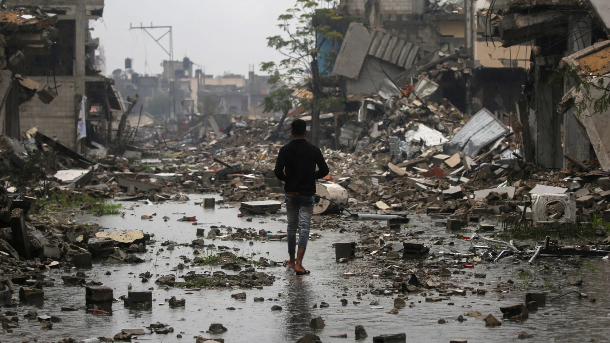 A Palestinian youth walks in the rain amidst destroyed buildings in Bureij in the central Gaza Strip on 23 January 2025 (AFP/Eyad Baba)