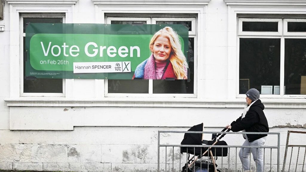 A woman walks past a sign for the Green party along a street in Denton, Greater Manchester, north-west England on February 25, 2026, the day before the crucial Gorton and Denton by-election. (AFP)