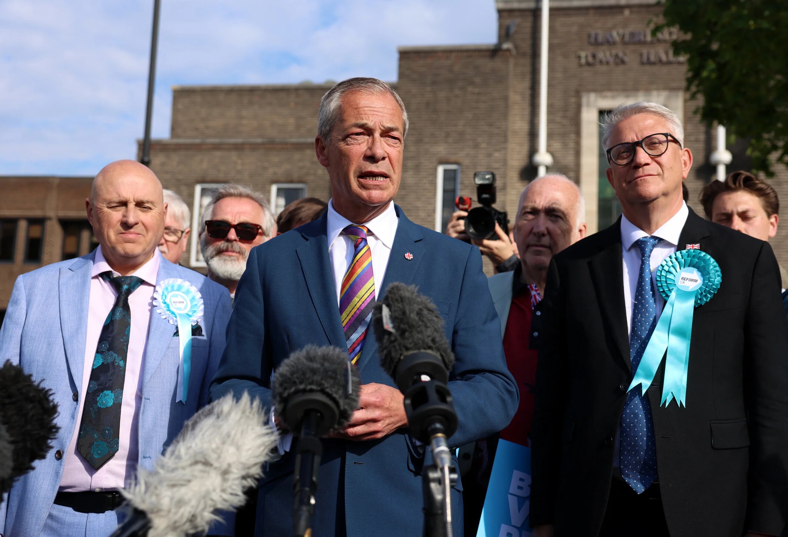 Reform UK leader Nigel Farage is pictured in a blue suit and a multi-coloured tie with microphones pointed at him as he speaks to the press