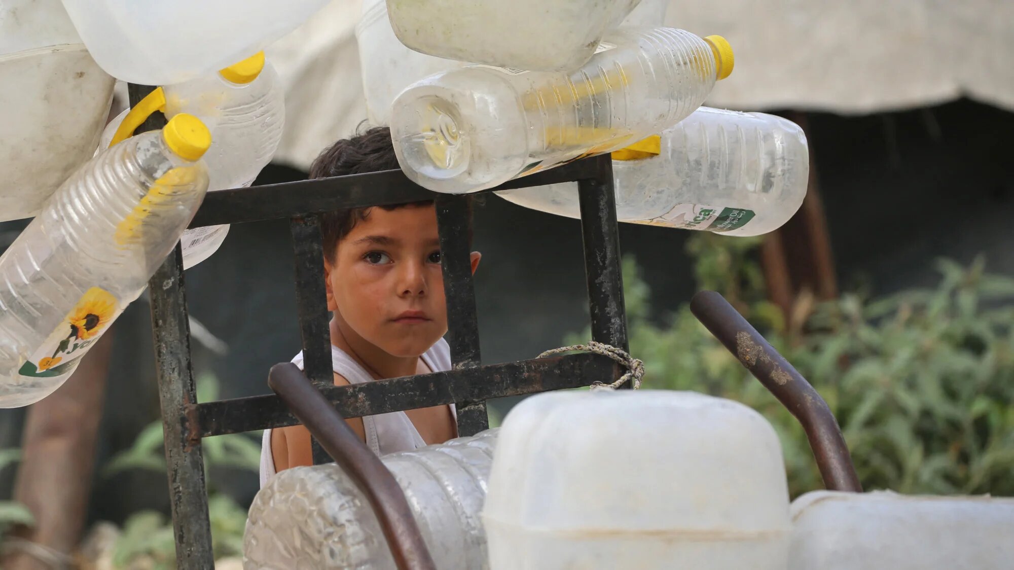 A Palestinian boy stands next to plastic containers on a trolley, as he and others collect portable water for their displaced family living in shelters in the Bureij refugee camp, in the central Gaza Strip on 18 May 2026 (Eyad Baba/AFP)