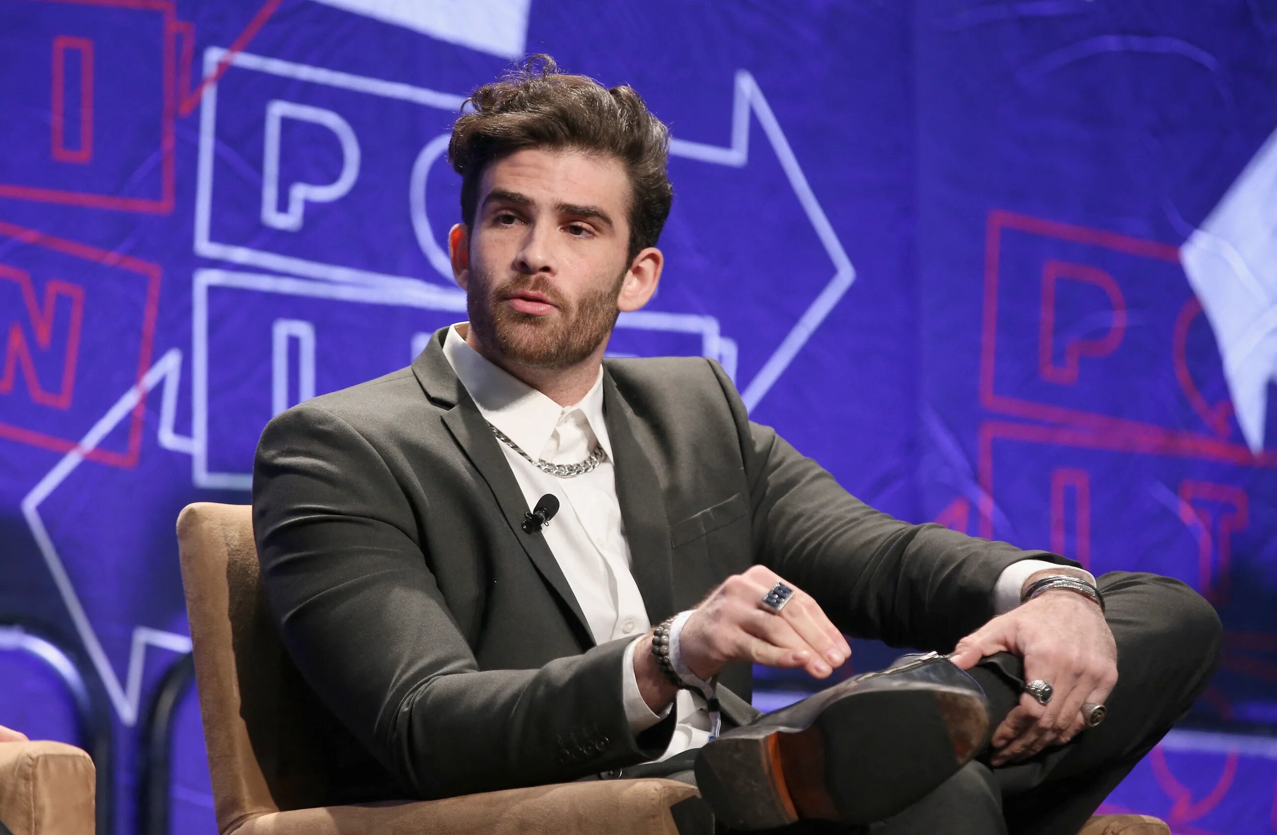 Hasan Piker in a white shirt and grey suit sitting on stage looking to one side
