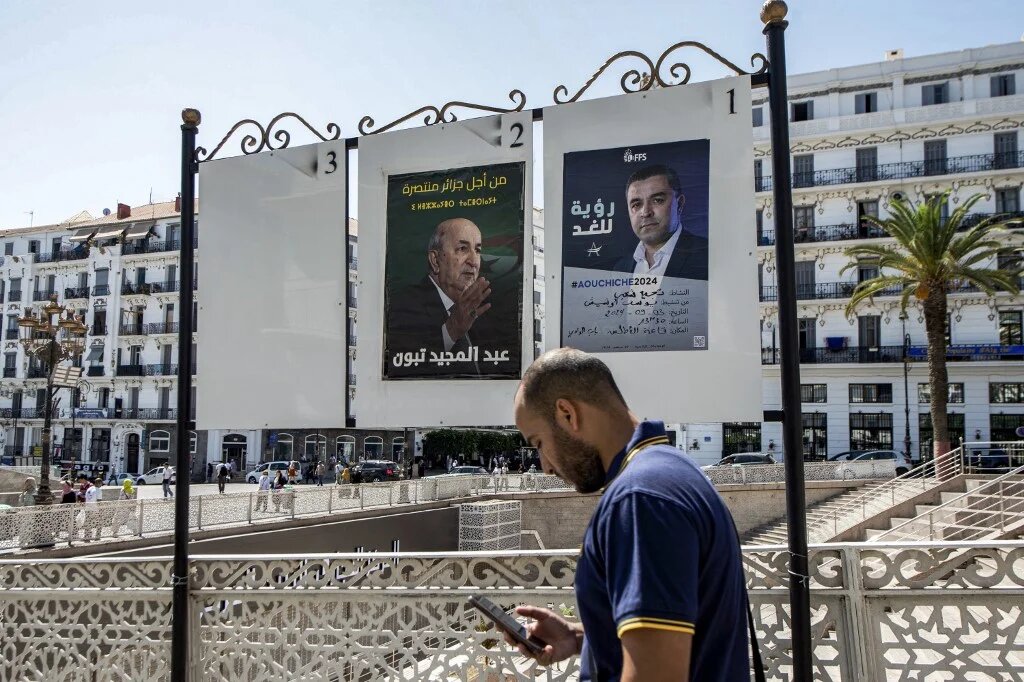 A man browses his phone as he walks past posters for presidential election candidates Abdelmadjid Tebboune (left) and the Socialist Forces Front's Youcef Aouchiche in Algiers, in September 2024 (AFP)