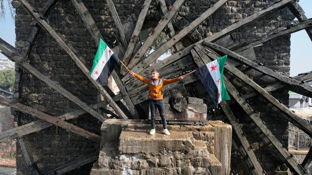A boy waves the opposition Syrian flag as he stands in one of the water wheels, or norias, in Hama on 6 December (Omar Haj Kadour/AFP)