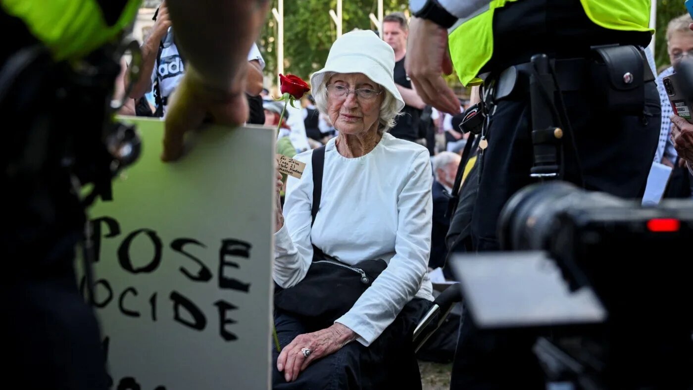 An 89-year-old protester named La holds a rose, as a police officer holds her placard reading "I oppose genocide, I support Palestine Action", during a rally organised by Defend Our Juries on 8 August 2025 (Jaimi Joy/Reuters)