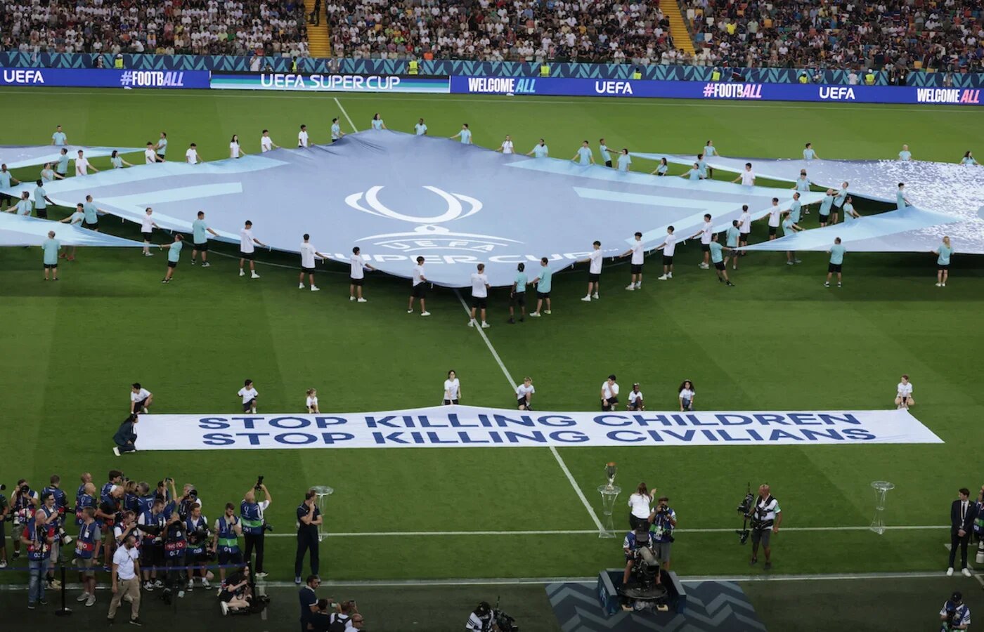 Refugee children display 'Stop killing children stop killing civilians' banner on the pitch ahead of the 2025 Uefea Super Cup final between Paris Saint-Germain and Tottenham Hotspur FC at the Friuli stadium, Udine, on 13 August 2025 (Remo Casilli/Reuters)