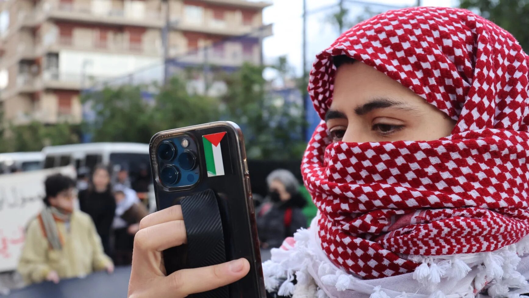 An activist wearing a chequered keffiyeh takes pictures with a smart phone bearing a Palestinian flag sticker as demonstrators block the road leading to the Egyptian embassy in the Lebanese capital Beirut in protest against the closure of the Rafah crossing between Egypt and the Gaza Strip, on February 19, 2024. Egypt, which controls the Rafah border crossing, has repeatedly warned against any "forced displacement" of Palestinians from Gaza into the Sinai desert.