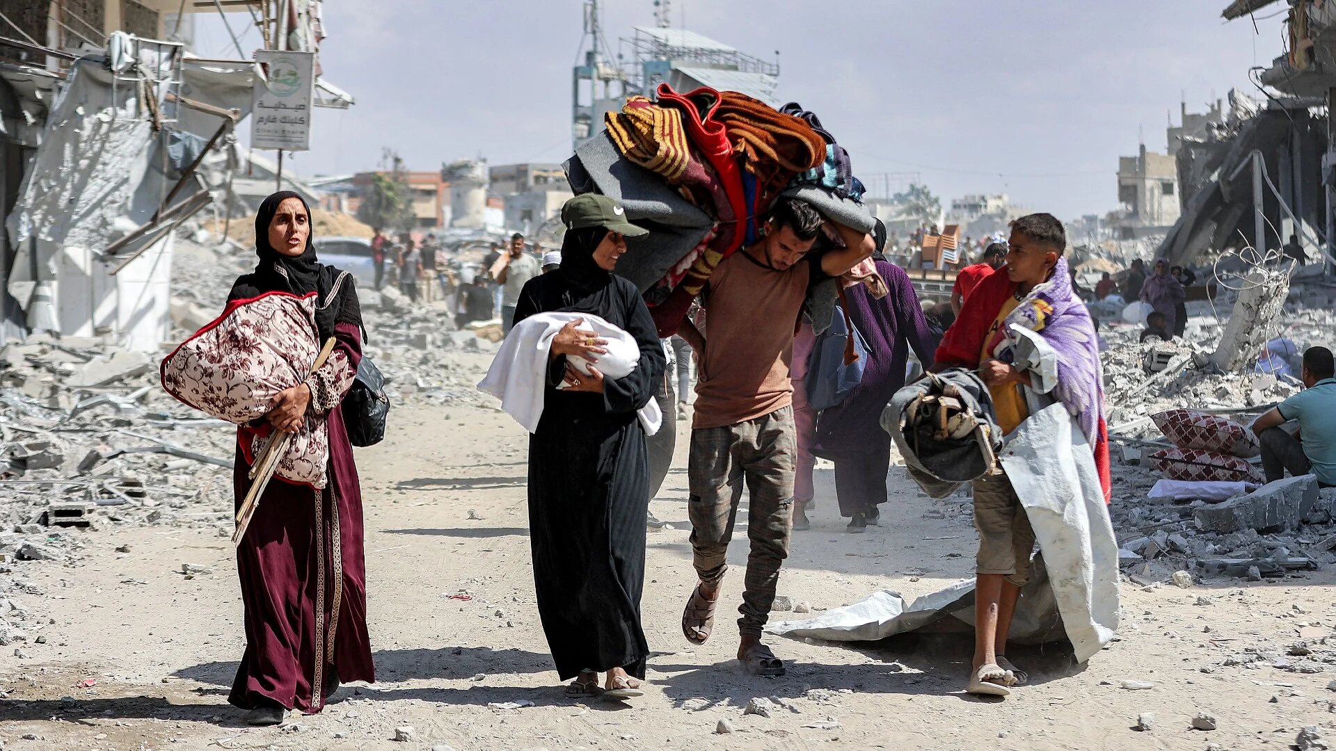 A woman carrying a child walks with other Palestinians past the rubble of destroyed buildings in the centre of Khan Younis in the southern Gaza Strip on 10 October 2025 (Omar al-Qattaa/AFP)