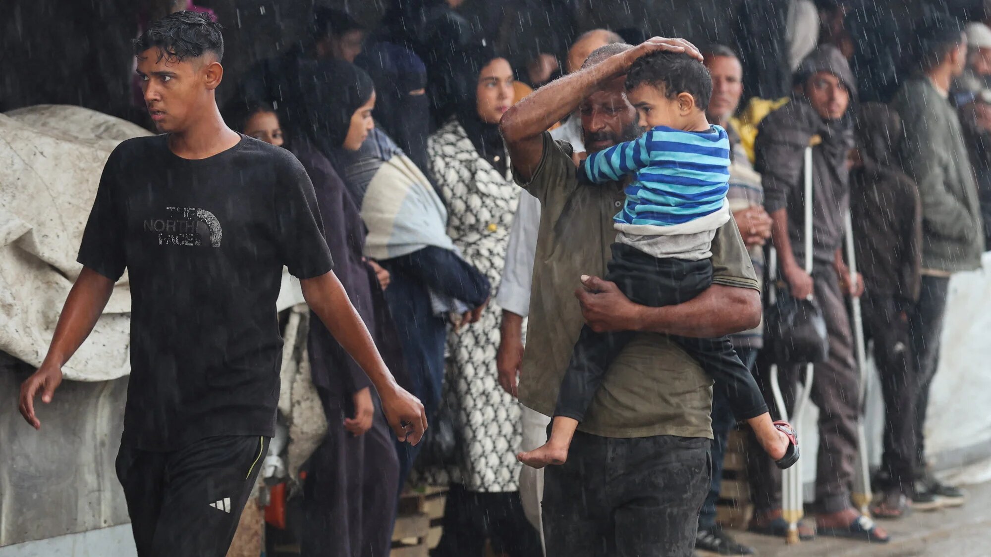A Palestinian shields a child from the rain as they rush to shelter during a downpour in Khan Younis, in the southern Gaza Strip, 25 November 2025 (Reuters)