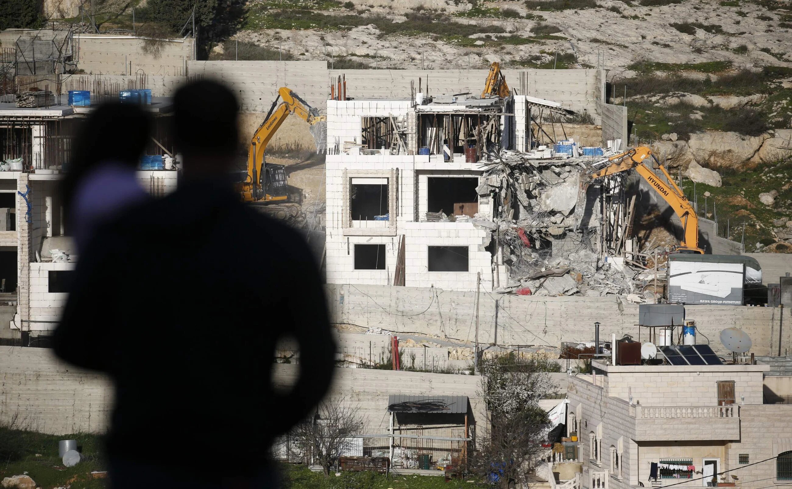 A Palestinian holds a child as he watches Israeli hydraulic shovels demolishing a Palestinian building, north of the occupied West Bank city of Hebron on 14 February 2018 (AFP)