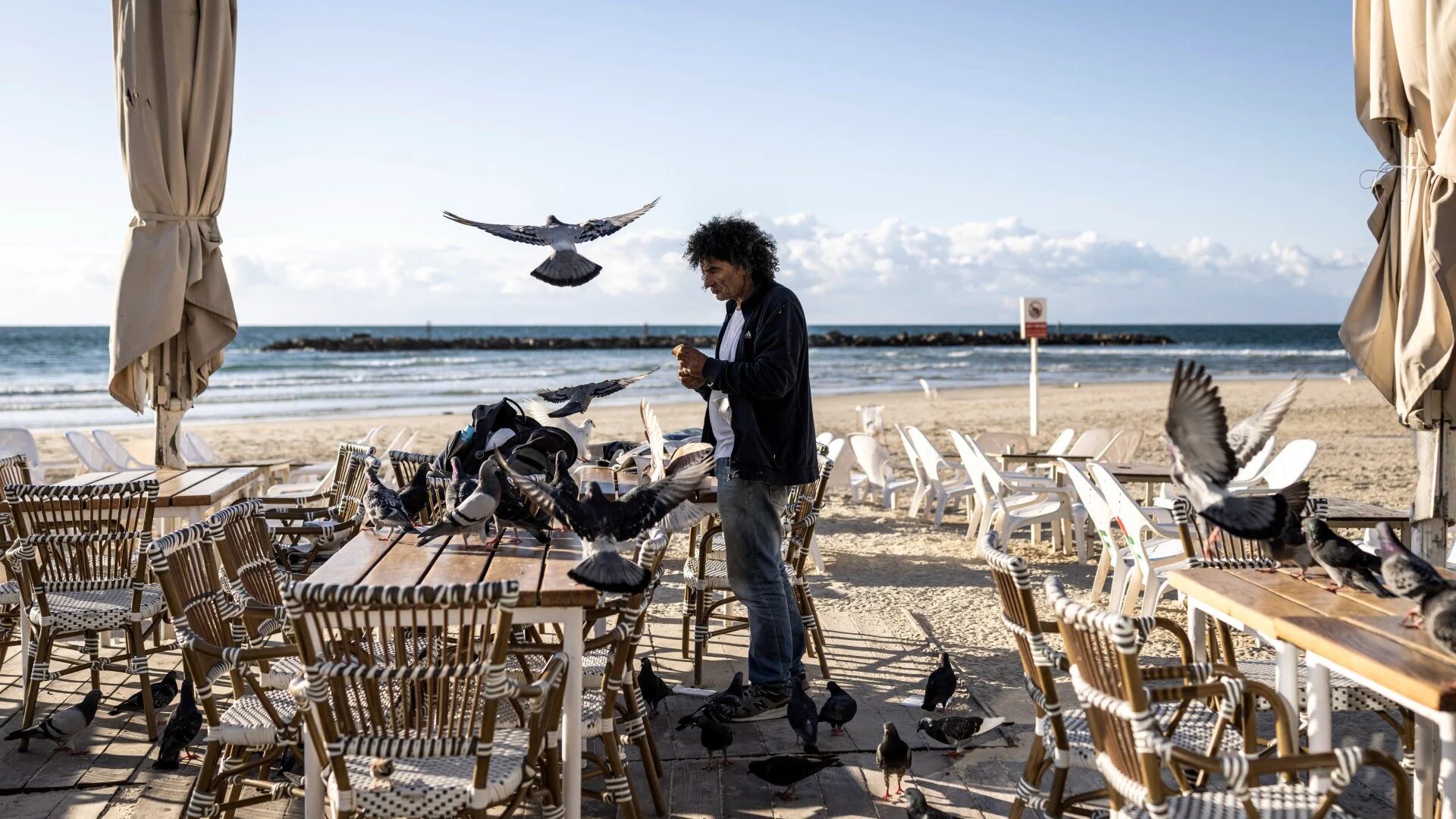 A man feeds pigeons along an empty beach in Tel Aviv on 28 February 2026 (John Wessels/AFP)
