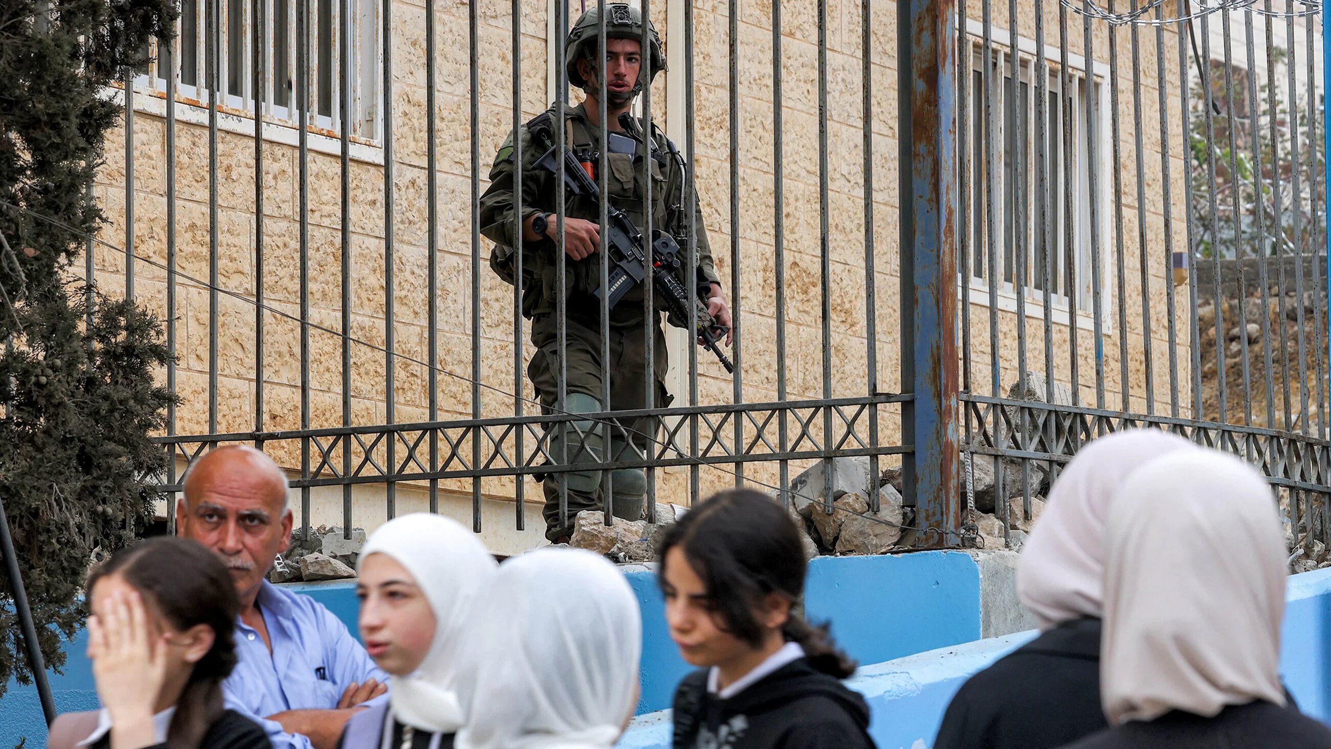 An Israeli army soldier stands from behind a fence as Palestinian schoolgirls make their way to school on the first day of term in Hebron, occupied West Bank on 8 September, 2025 (AFP/Hazem Bader)