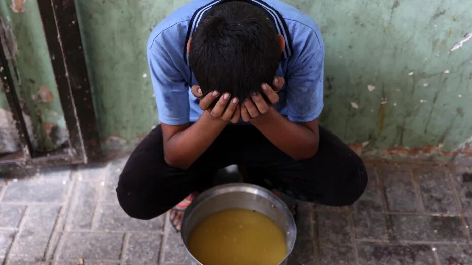 A displaced Palestinian child sits next to a pot of lentil soup that he received at a food distribution point in Gaza City on 25 July 2025 (Omar al-Qattaa/AFP)