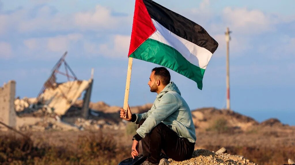 A man holds a Palestinian flag as he watches displaced Palestinians cross the Netzarim Corridor from southern Gaza into Gaza City on 27 January 2025 (Omar al-Qattaa/AFP)