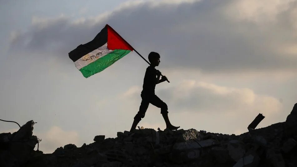 A displaced child waves a Palestinian national flag as he walks on the rubble of a building in Gaza’s Bureij refugee camp, on 22 September 2025 (Eyad Baba/AFP)