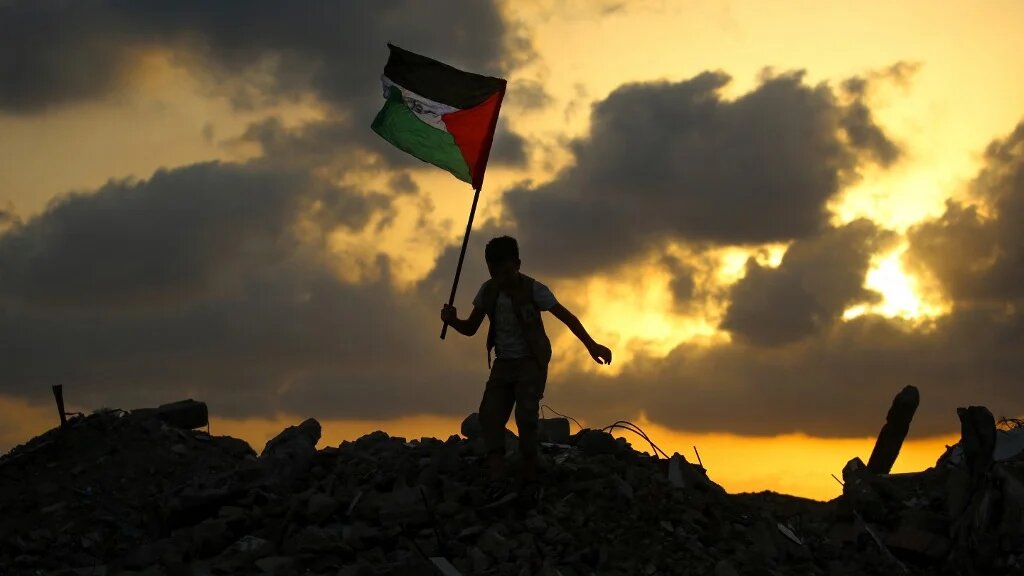 A displaced child waves the Palestinian national flag atop the rubble of a destroyed building in Gaza’s Bureij refugee camp on 22 September 2025 (Eyad Baba/AFP)