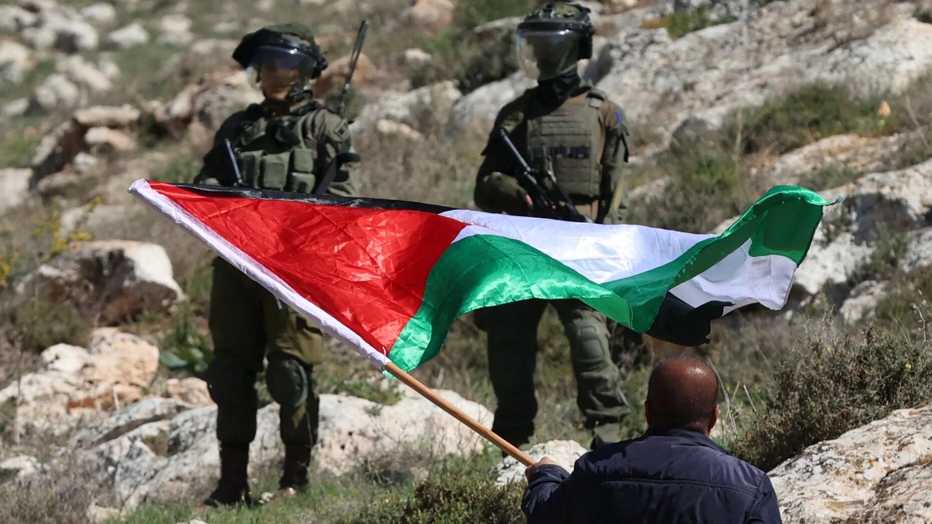 A Palestinian demonstrator facing Israeli troops waves a national flag during a protest against the establishment of Israeli outposts, near the occupied-West Bank city of Nablus, on 10 March, 2023 (AFP)