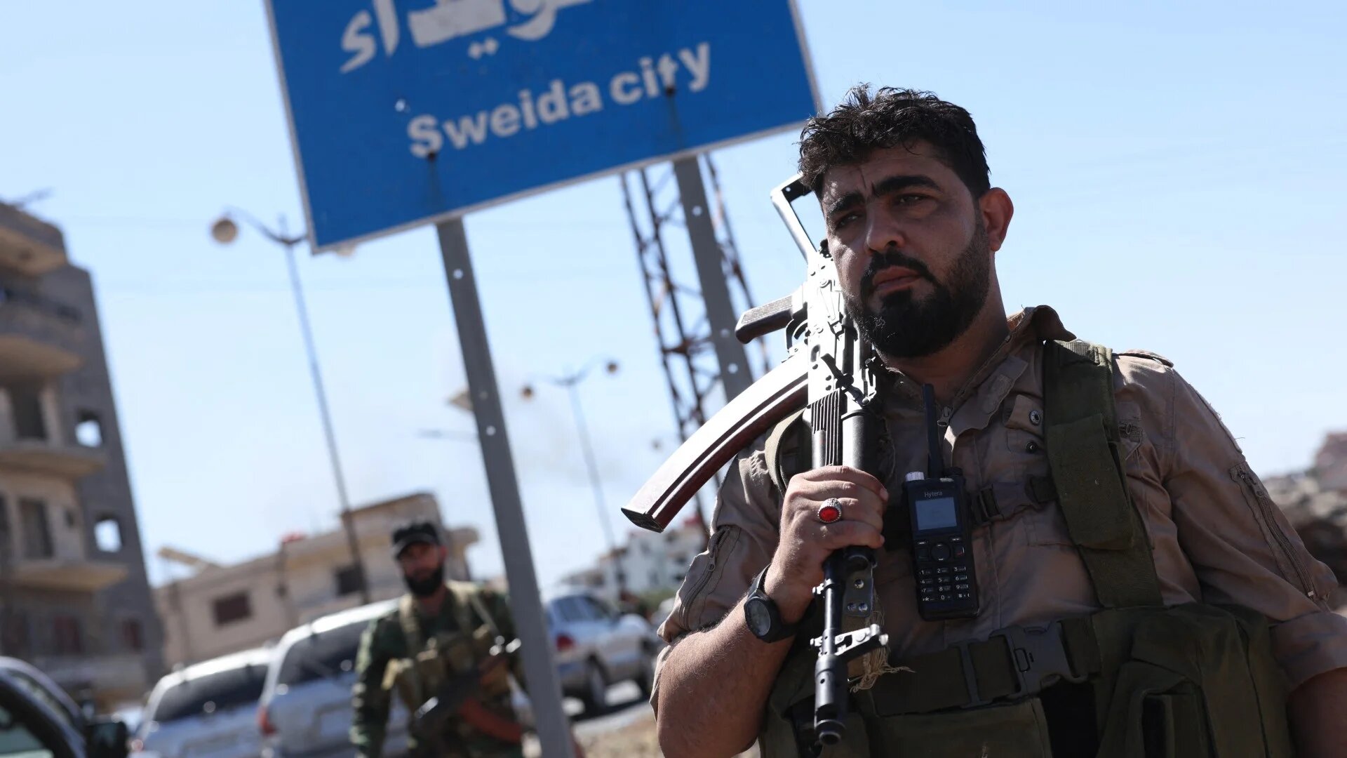 A member of Syria's security forces stands under a sign in the predominantly Druze city of Sweida on 15 July (Bakr Alkasem/AFP)