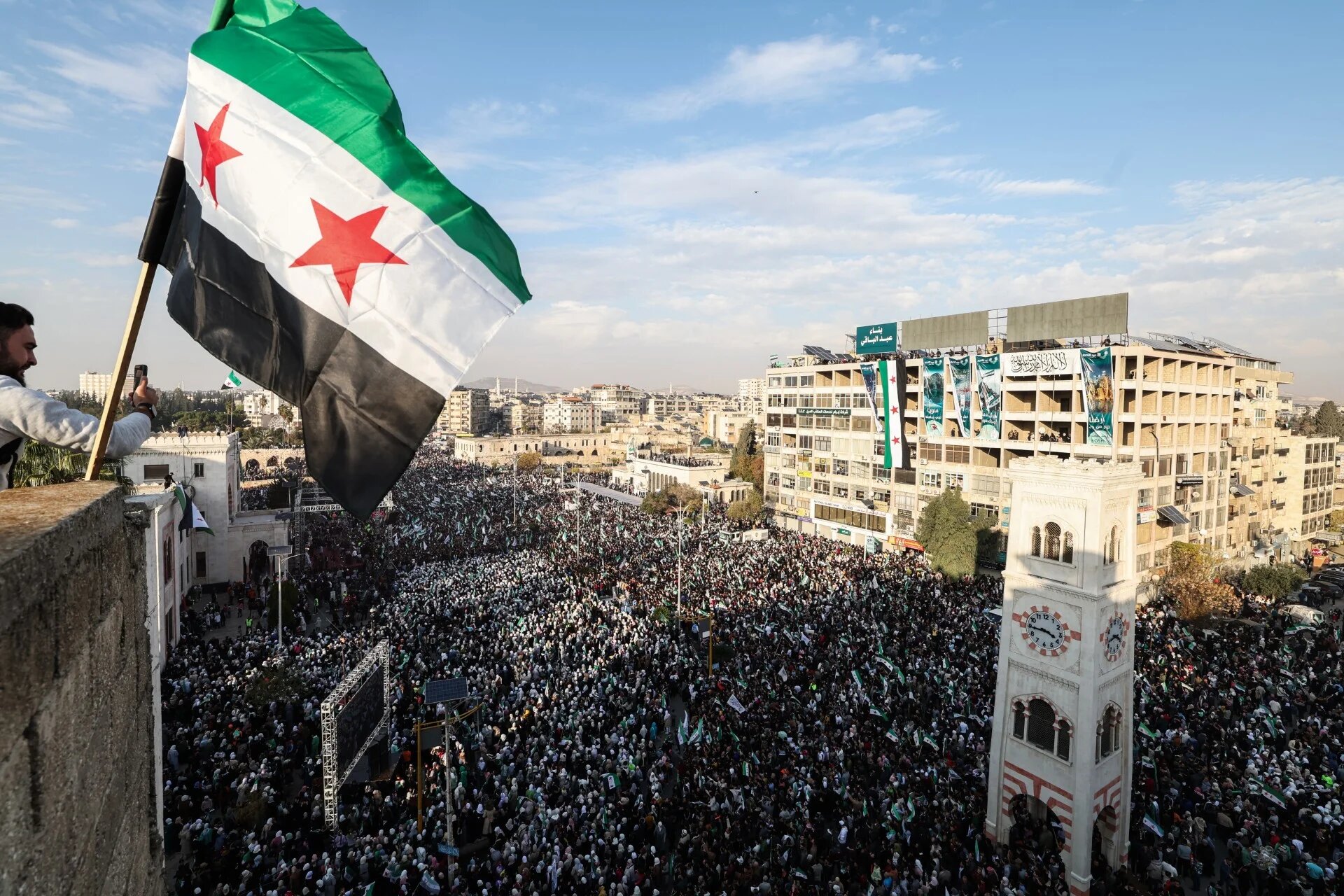 A Syrian flag flutters above crowds gathering during celebrations marking one year since a lightning offensive that eventually toppled the country's longtime ruler, in central Hama on 5 December 2025 (Omar Haj Kadour/AFP)