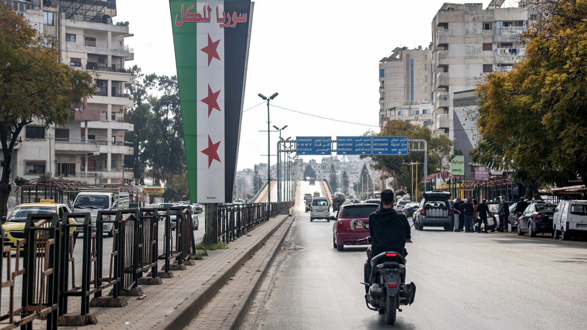 Motorists drive past a large sign depicting the new Syrian flag, with a slogan in Arabic reading "Syria for all", in the western city of Latakia on 9 March 2025 (AFP/Omar Haj Kadour)
